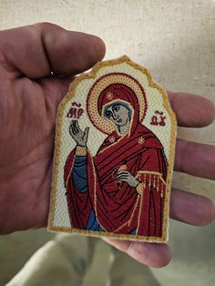 a hand holding a small embroidered icon of the virgin mary in red and gold colors