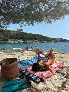 a woman laying on top of a towel next to a tree near the ocean and people swimming in the water