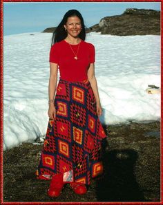 a woman standing in the snow wearing a red shirt and colorful skirt with an orange necklace