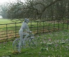 a statue of a man riding a bike in the grass near a fence and flowers