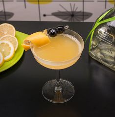 an alcoholic drink in a coupe glass on a table with lemons and a bottle