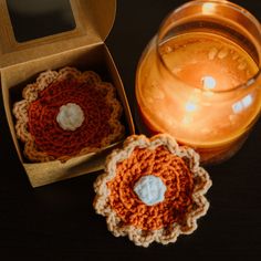 two crocheted coasters sitting next to a candle on a table with an open box