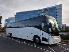 a large white bus parked in a parking lot next to a tall building with glass windows