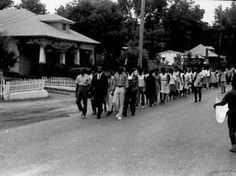 black and white photograph of people walking down the street in front of houses with picket fences