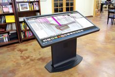 a computer screen in a library with bookshelves and tables behind it on the floor