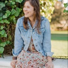 a woman sitting on a ledge wearing a dress and jean jacket, smiling at the camera