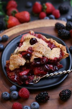 a slice of berry pie on a black plate with berries and raspberries around it