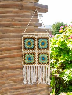 a crocheted wall hanging on the side of a wooden building with flowers in the background