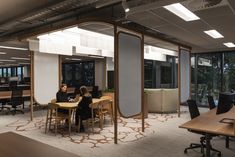 two people sitting at a table in an office setting with large windows and wooden tables