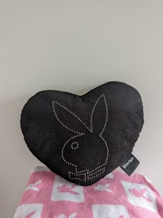 a black heart shaped pillow sitting on top of a pink and white bed sheet with an embroidered bunny face