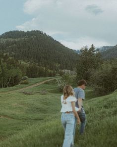 a man and woman walking through a lush green field with mountains in the back ground