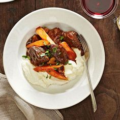 a white plate topped with meat and mashed potatoes next to a glass of wine