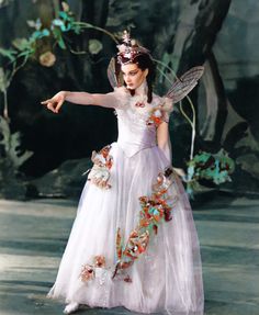 a woman in a white dress with flowers on her head and wings, pointing at something