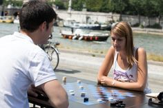 a man and woman sitting at a table playing chess in front of the water with bicycles