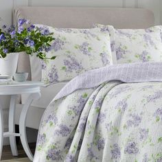 a bed with purple and green flowers on the comforter next to a white table