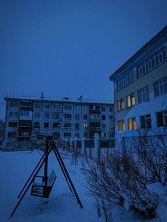 a swing set in the snow next to some apartment buildings at night with lights on