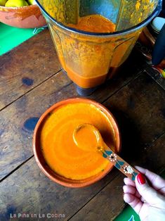 a person holding a spoon over a bowl of orange liquid next to a blender