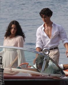 a man and woman standing next to a boat in the water with one holding on to the steering wheel