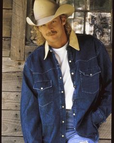 a man wearing a cowboy hat and jeans leaning against a wooden wall with his hands in his pockets