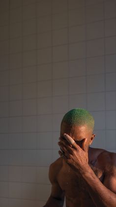 a shirtless man with green hair covering his face in front of a white tiled wall