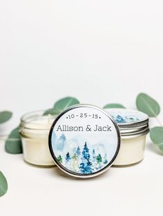 two small jars with personalized labels sitting next to some green leaves on a white surface