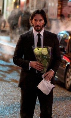 a man in a suit and tie holding flowers on the side of the road with cars behind him