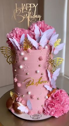 a pink birthday cake with gold butterflies and flowers on the top is sitting on a table