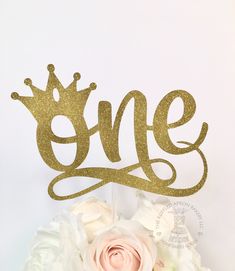 a cake topper that says one with a crown on it and flowers in the foreground