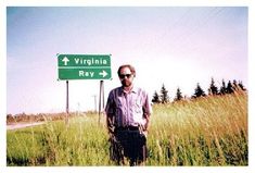 a man standing in tall grass next to a road sign that says virginia and ray