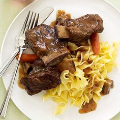 a white plate topped with meat and pasta next to a fork on a tablecloth