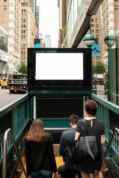 three people are walking up the stairs towards an empty billboard in a city with tall buildings