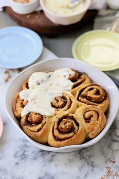 cinnamon rolls with icing in a white bowl on a marble countertop next to plates