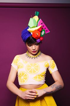 a woman in a yellow skirt and crop top with fruit on her head standing against a purple wall