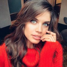 a woman with blue eyes wearing a red sweater and holding her hand to her face