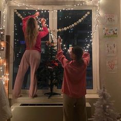 two people standing in front of a window with christmas lights hanging from the windowsill