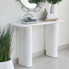 a white console table with two plants and a round mirror on the wall behind it