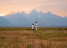 two people standing in a field with mountains in the backgrounnd and one person holding hands