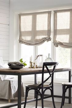 a dining room table and chairs in front of two windows with roman shades on them