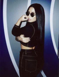 a woman with long black hair and sunglasses posing for a photo in front of a wall