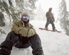 two snowboarders are sitting in the snow with their boards on and one is wearing goggles