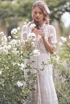 a woman in a white dress is standing among flowers and plants with her hands on her chest