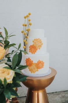 a three tiered white cake with orange flowers on top and gold stand next to it