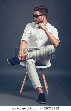 a man in white shirt and pants sitting on a chair with his hand under his chin