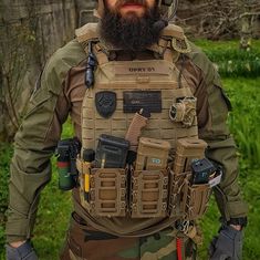 a bearded man wearing an army uniform and helmet with tools on his back, standing in the grass