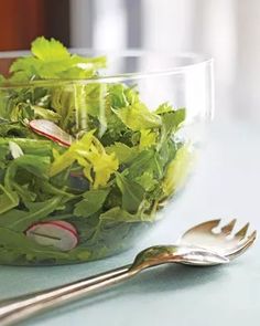 a salad in a glass bowl next to a fork and spoon on a blue table