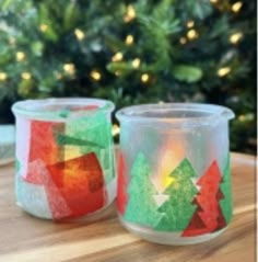 two glass candles sitting on top of a wooden table next to a christmas tree in the background