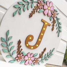 a sign with the letter j on it is decorated with flowers and leaves in gold