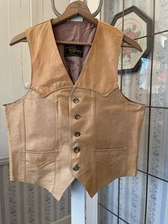 "This vest is made of very soft leather in two tones of light tan brown. It has the original buttons in the front and two front pockets. The front and back yokes and pockets are trimmed with matching braided leather. The vest is fully lined with light brown acetate lining. The label reads \"Le Cuir Niko Leather.\" The measurements, taken with the vest lying flat, are: shoulder to shoulder, 13 1/2 inches; armpit to armpit, 22 inches; length, 25 inches in front and 21 inches in back; bottom edge, 22 inches. Please note that there are some spots on the right front; otherwise, the vest is in very good condition." Western Style Sleeveless Vest For Rodeo, Western Rodeo Vest, Brown Sleeveless Vest For Western-themed Events, Fitted Western Leather Vest, Leather Vest With Button Closure, Vintage Leather Vest Outerwear, Leather Sleeveless Vest With Button Closure, Sleeveless Leather Vest With Button Closure, Western Style Brown Vest For Western-themed Events