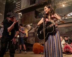 a woman singing into a microphone while holding an acoustic guitar in front of a man