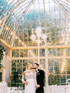 a bride and groom standing in front of a large glass building with lights hanging from the ceiling